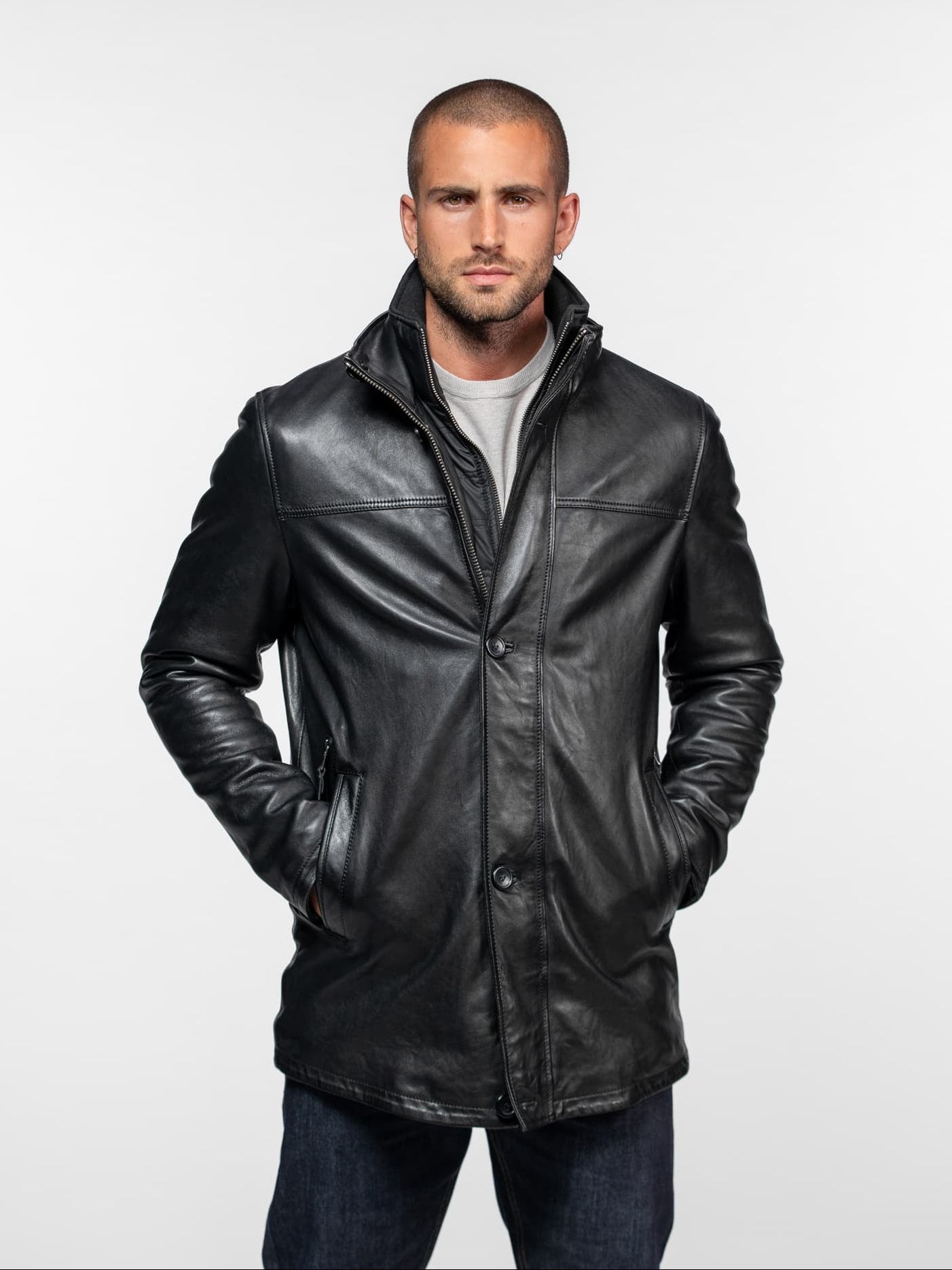 Man in a black leather jacket with hands in pockets, standing against a plain white background, exuding a confident demeanor.