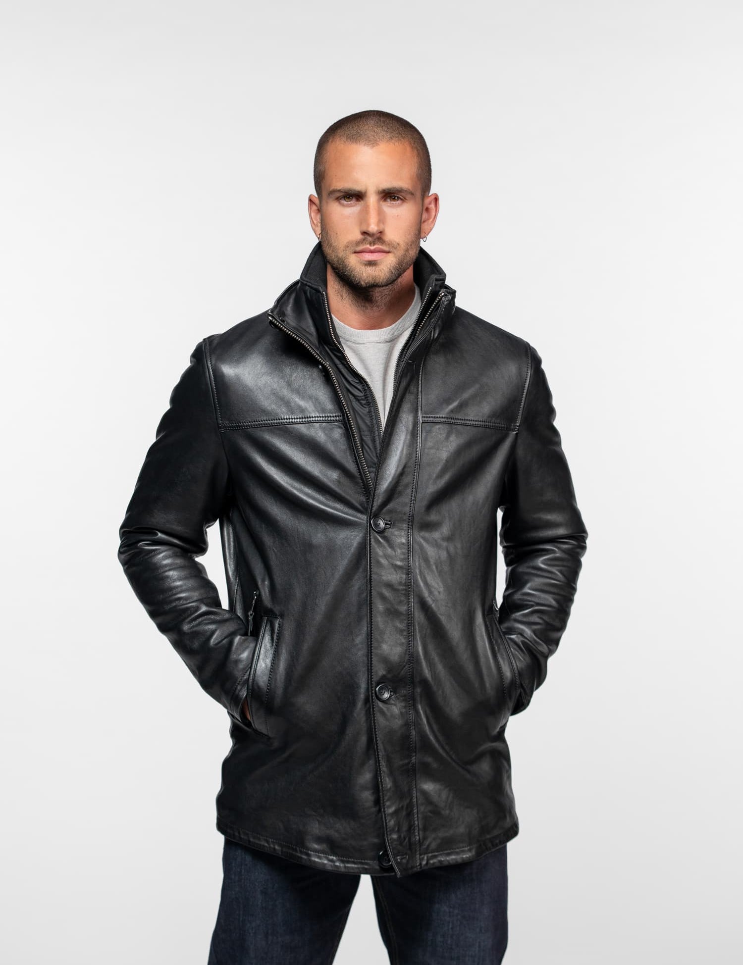 Man in a black leather jacket with hands in pockets, standing against a plain white background, exuding a confident demeanor.