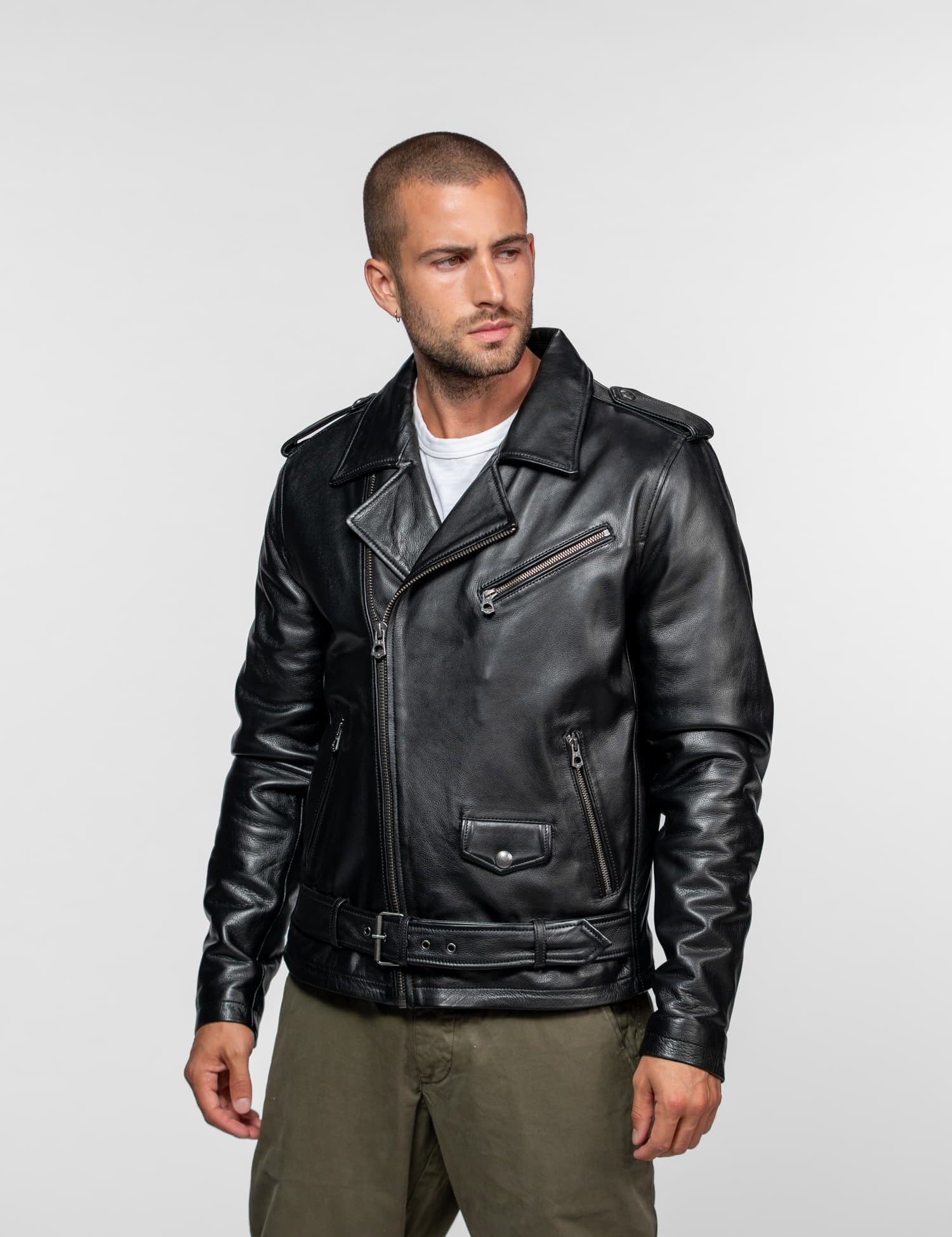 Man in a black leather jacket and khaki pants stands against a plain background, looking to the side with a serious expression.