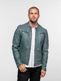 Man in a teal leather jacket and white shirt stands confidently against a plain background, hands in pockets, looking forward.
