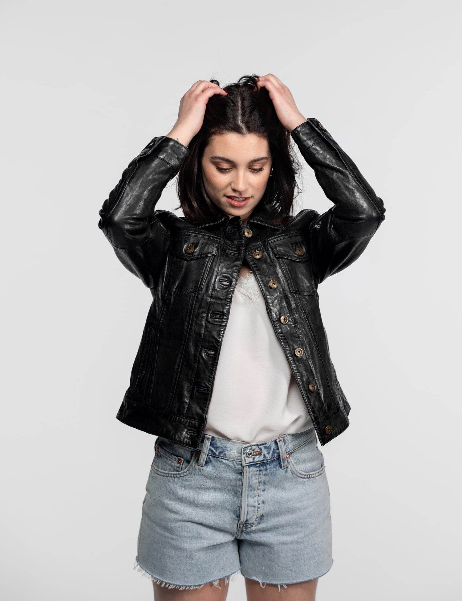 Woman in black leather jacket and denim shorts adjusts hair, standing against a plain background, looking down with a slight smile.