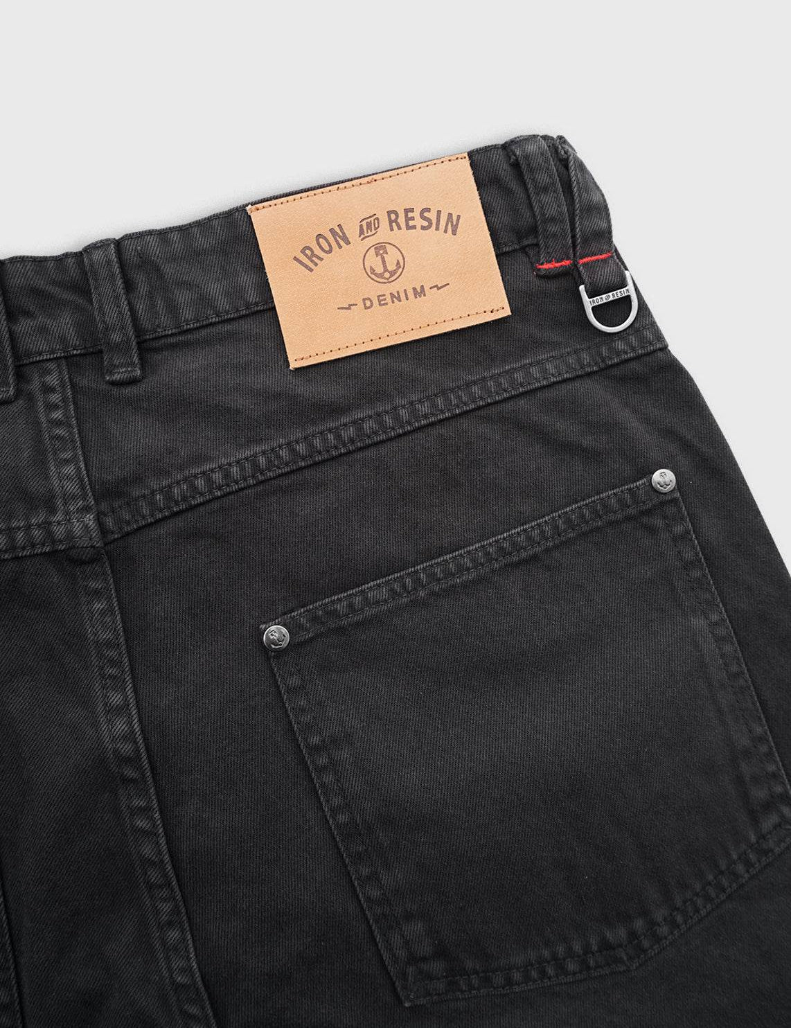Close-up of black denim jeans with a tan "Iron and Resin" label, featuring a back pocket and belt loop detail.