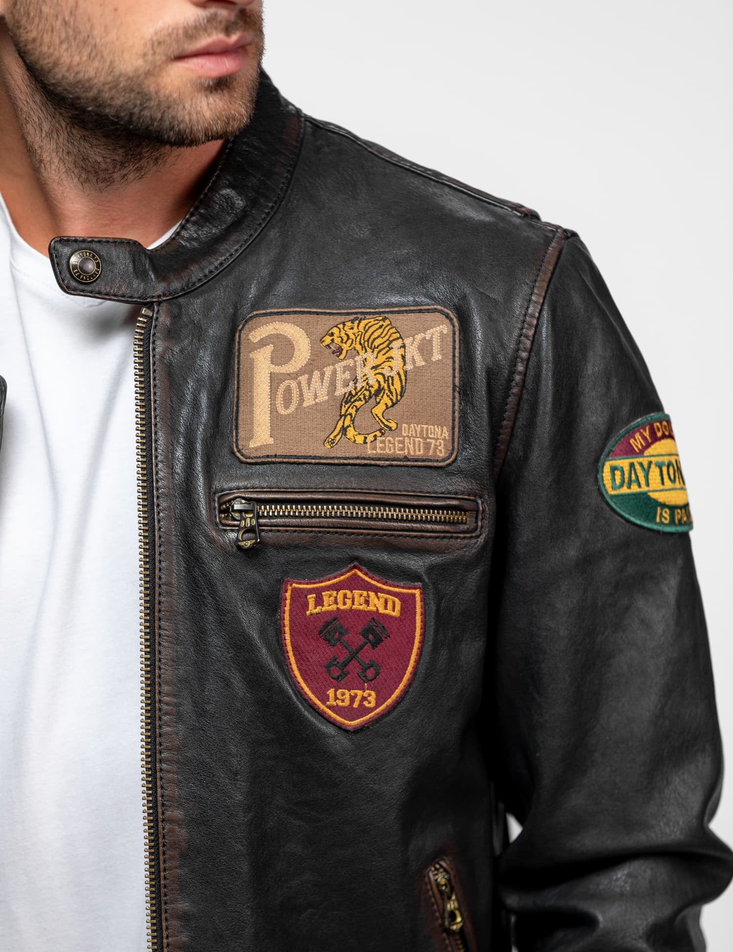 Man in a leather jacket with patches, including "Powerkit" and "Legend 1973," looking to the side against a plain background.