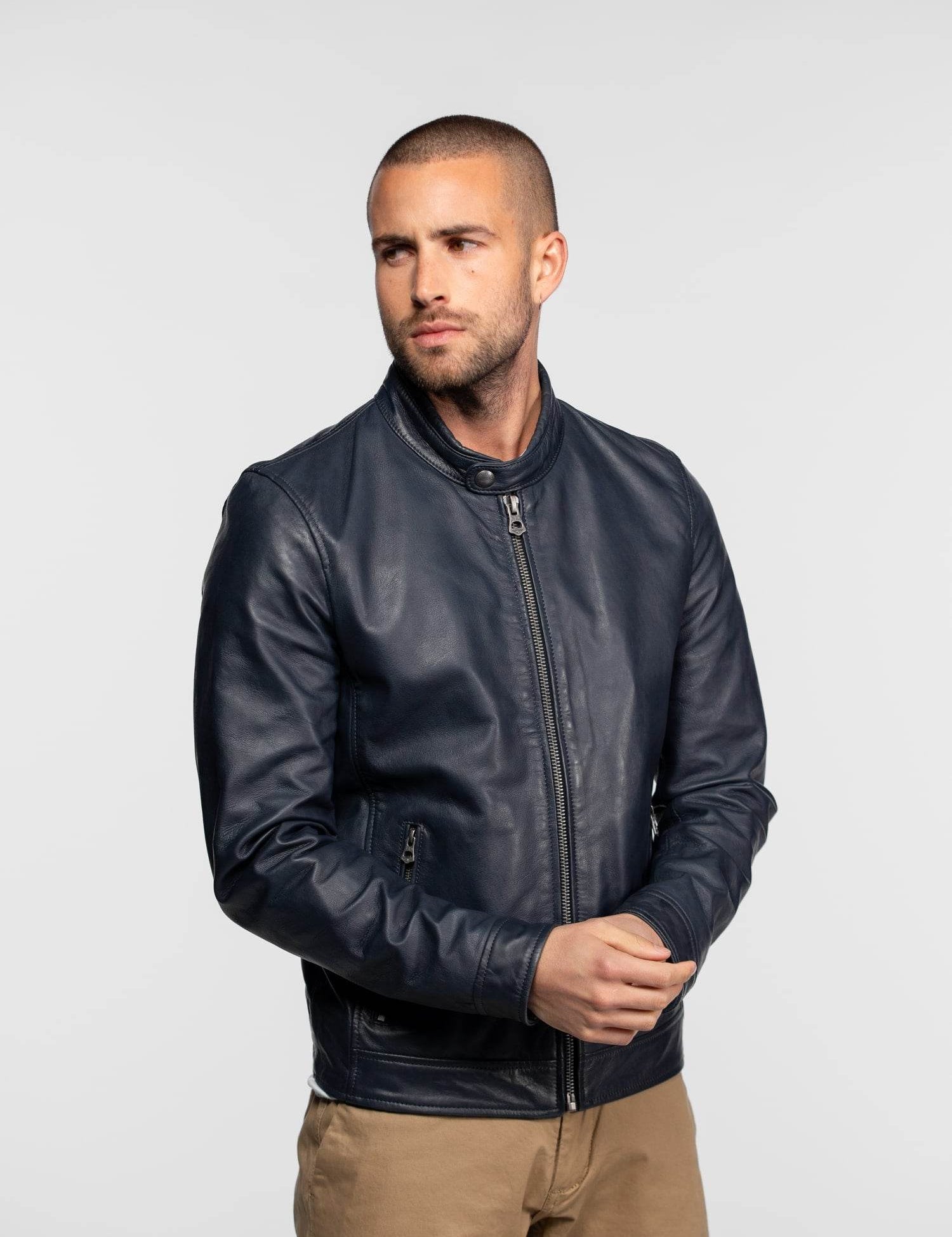 A man in a navy leather jacket and beige pants stands against a plain background, looking to the side with a thoughtful expression.
