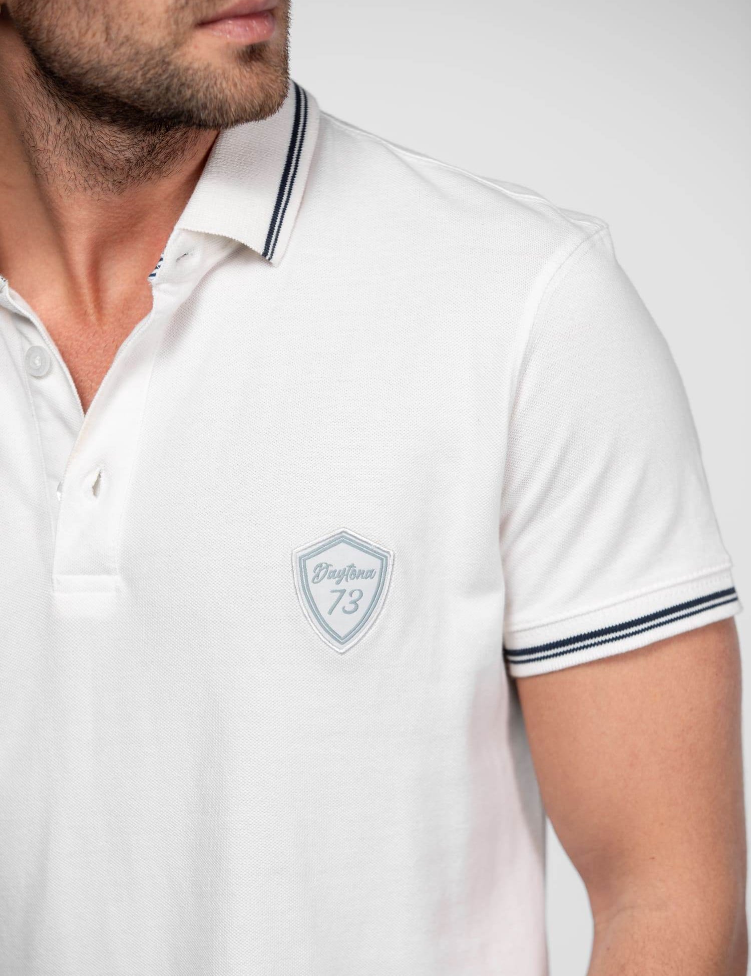 Man in a white polo shirt with "Daytona 73" logo, striped collar, and sleeves, looking to the side against a plain background.