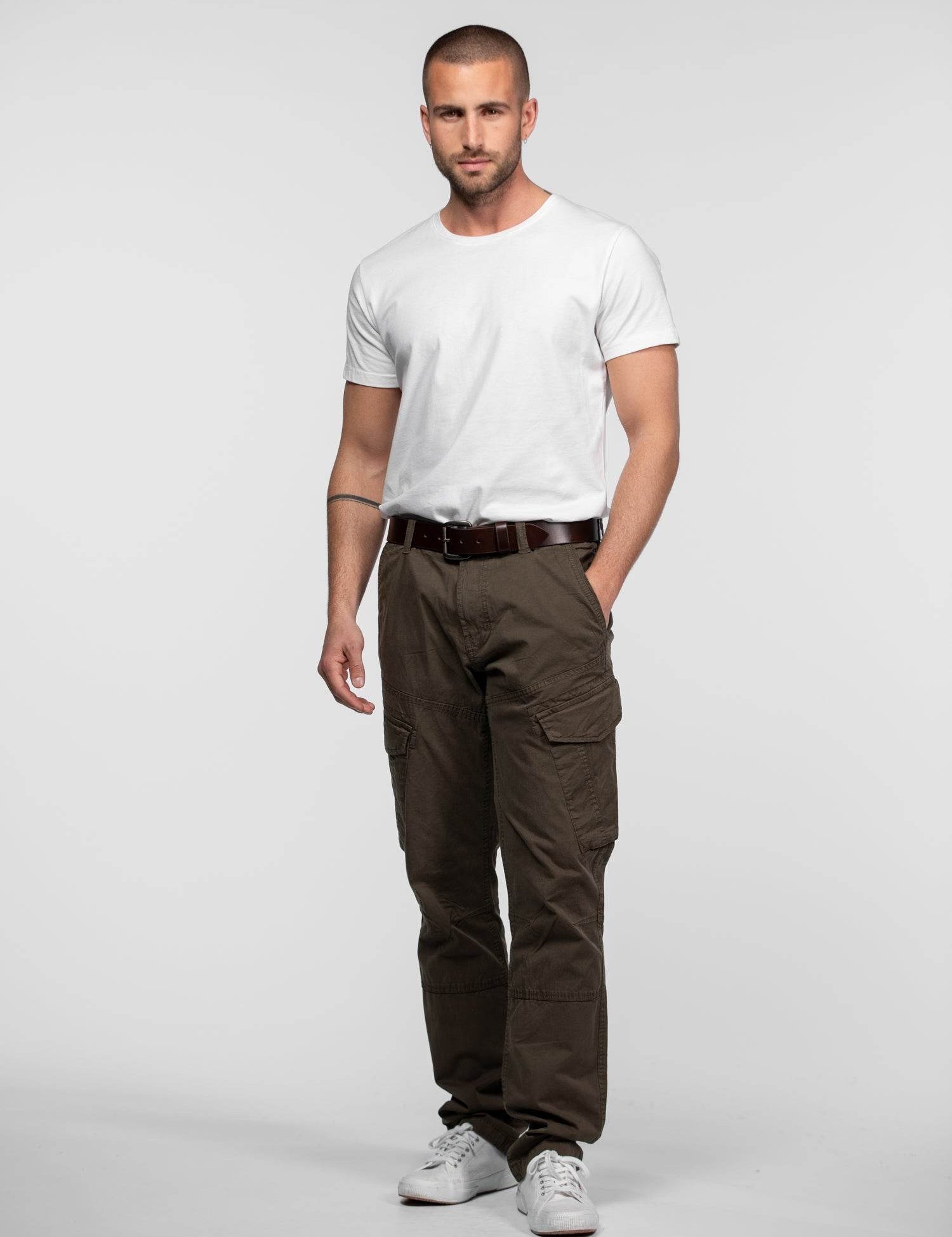 Man in a white t-shirt and brown cargo pants stands confidently against a plain background, hands in pockets, wearing sneakers.