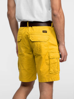 Man in white shirt and brown belt wearing bright yellow cargo shorts with multiple pockets, standing against a plain background.