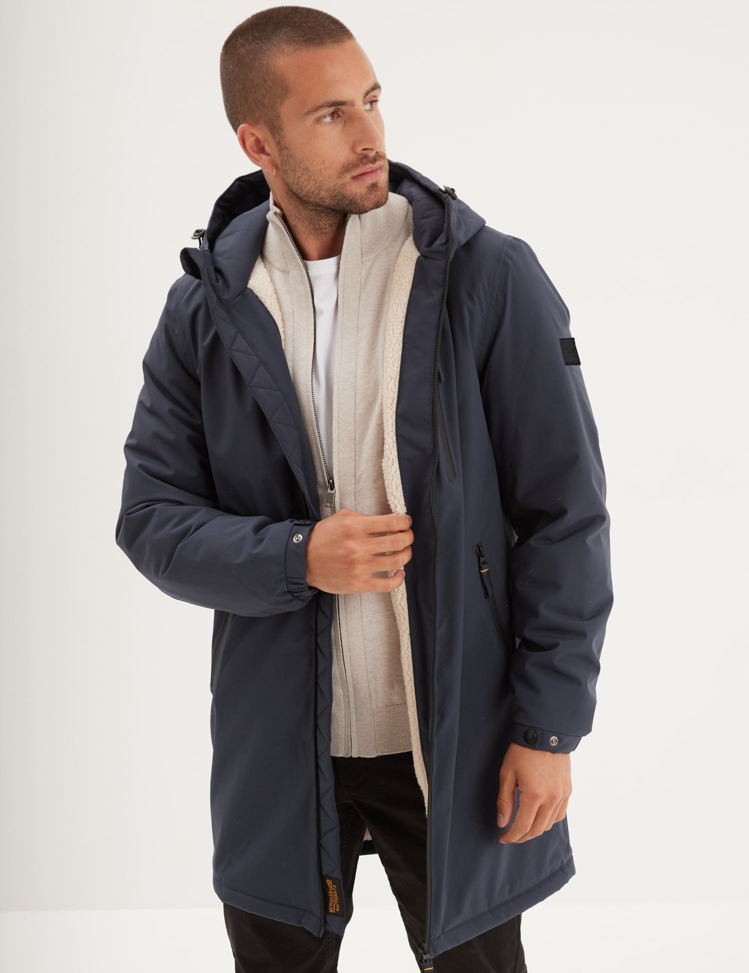 A man in a navy parka with a hood and beige lining, wearing a white shirt, looks to the side against a plain background.