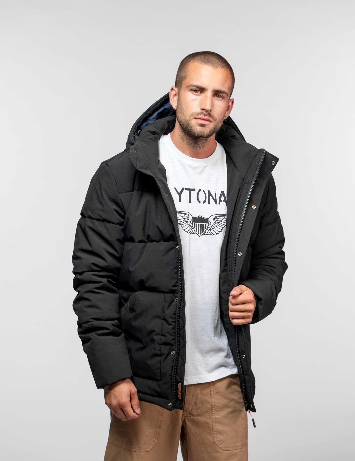 Man in a black puffer jacket over a white graphic tee and tan pants, standing against a plain background, looking at the camera.