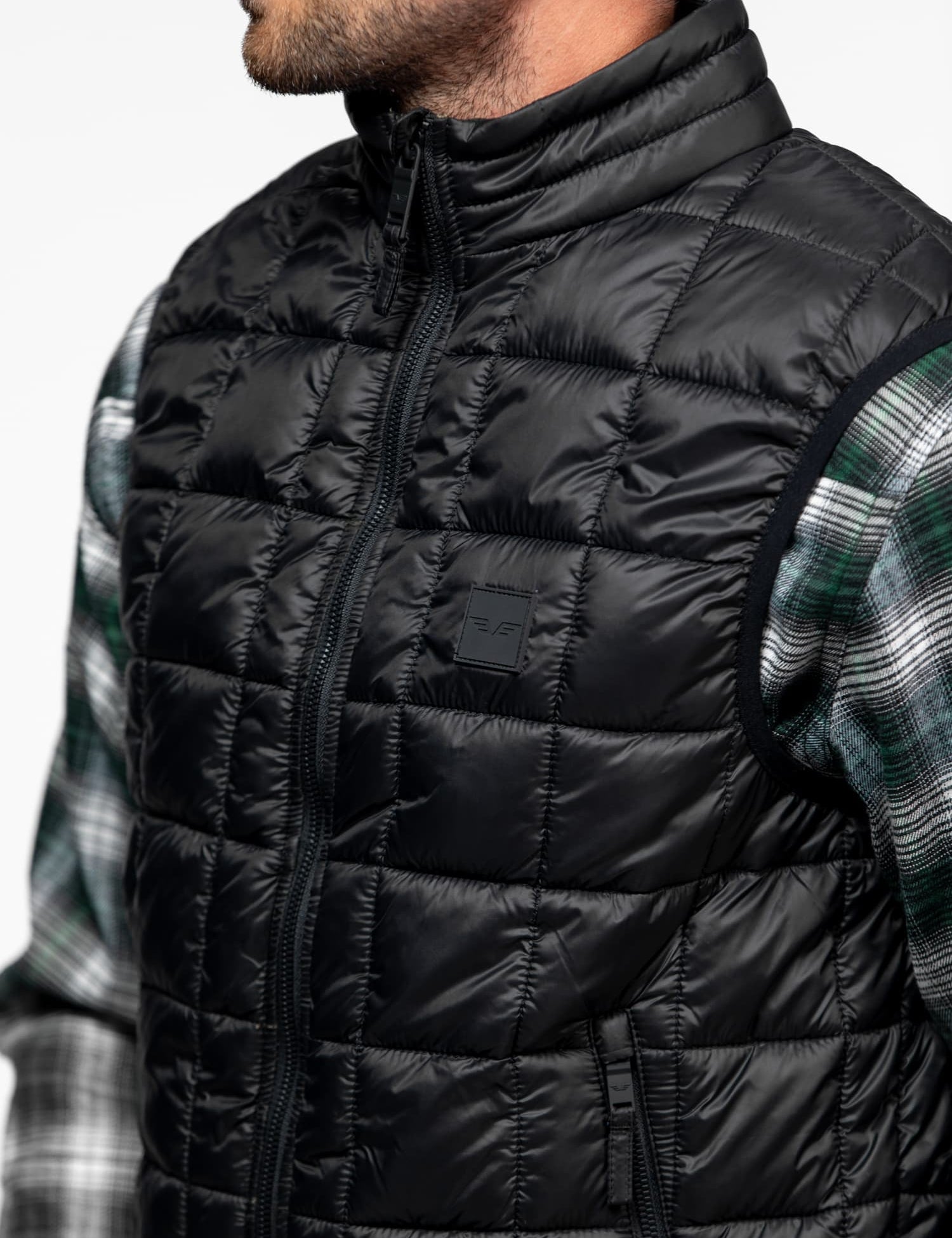 Man in a black quilted vest over a green plaid shirt, facing left. The vest features a high collar and visible stitching.