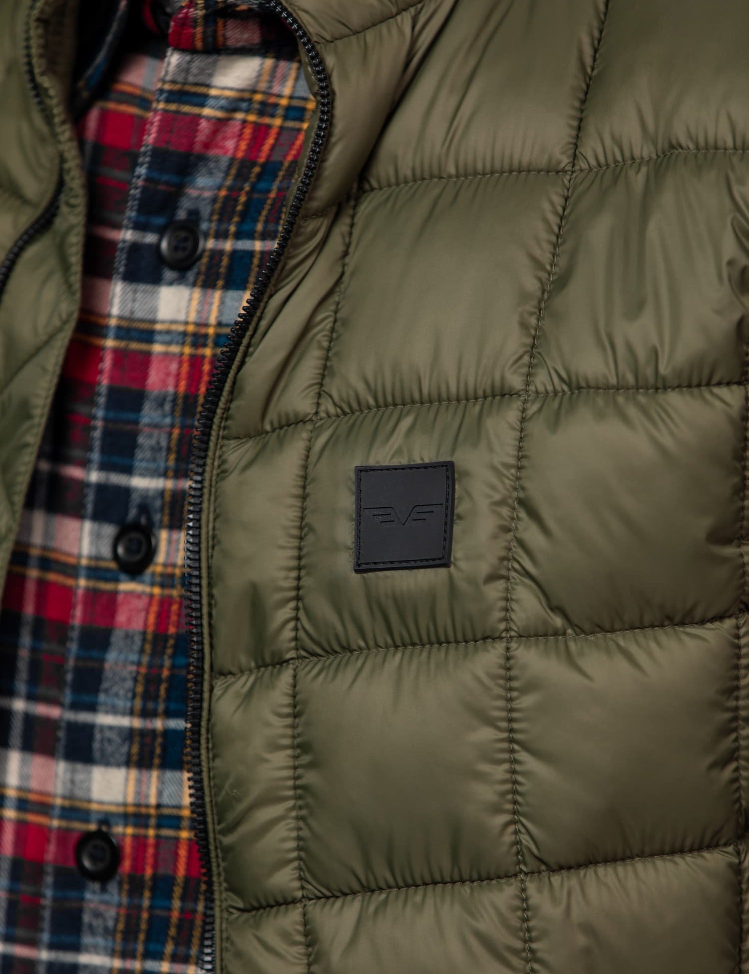 Close-up of a green quilted jacket with a logo, worn over a red and blue plaid shirt, highlighting texture and style.