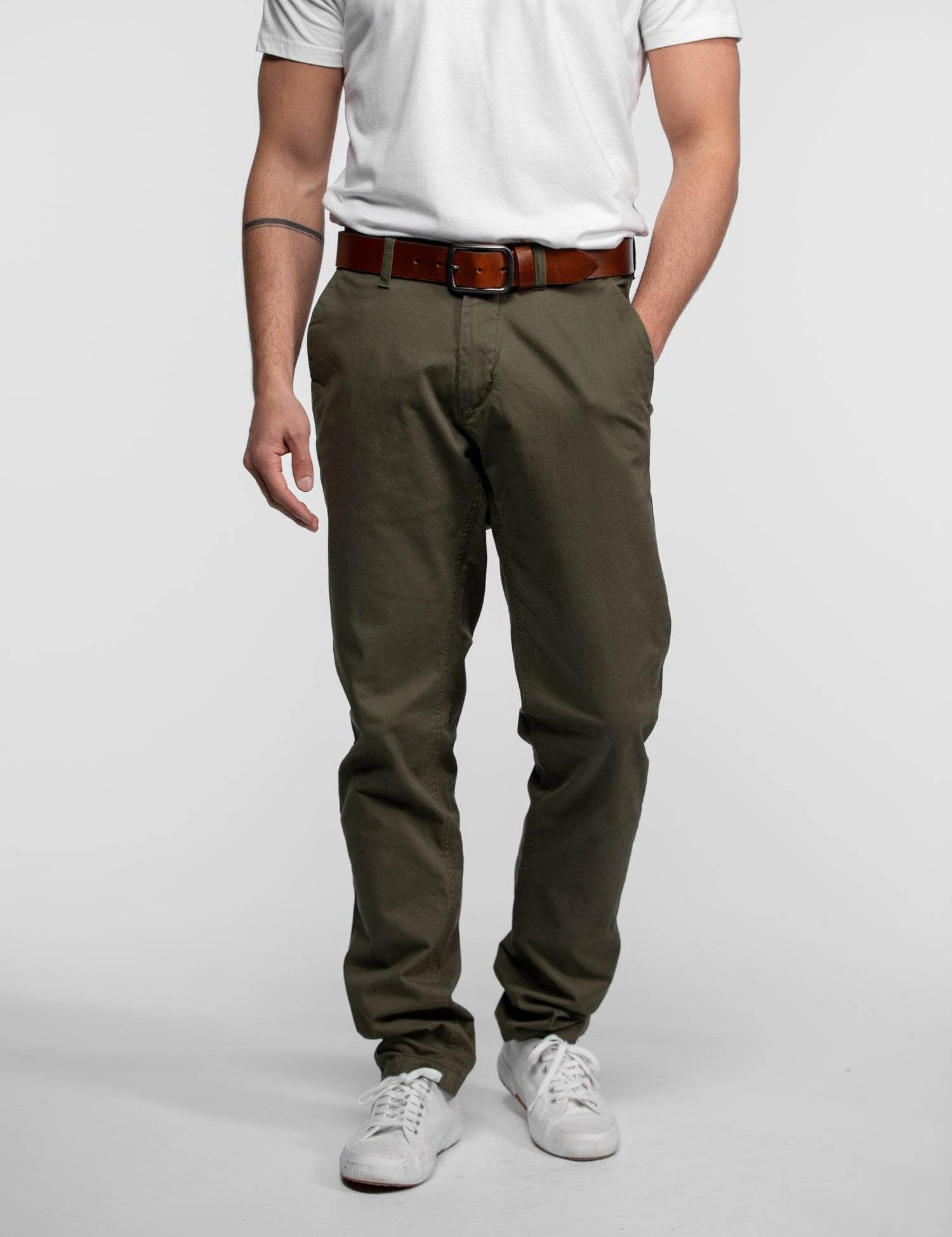 Man in white shirt, olive pants, brown belt, and white sneakers stands against a plain background, showcasing casual style.