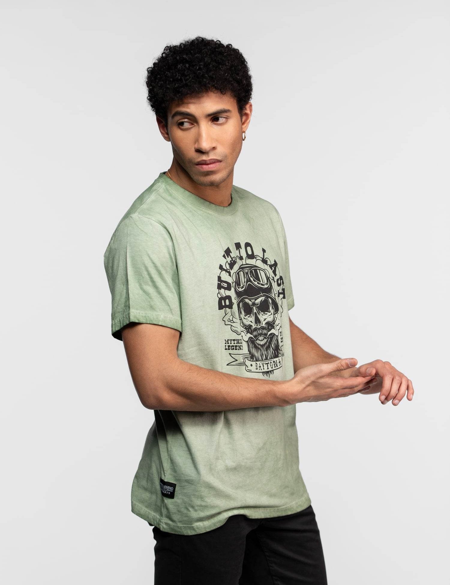Man in green "Built to Last" skull tee, looking sideways, with curly hair, stands against a plain background, hands clasped.