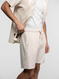 Man in a cream shirt and shorts adjusts his outfit, showcasing casual style. The focus is on the comfortable, neutral-toned fabric.
