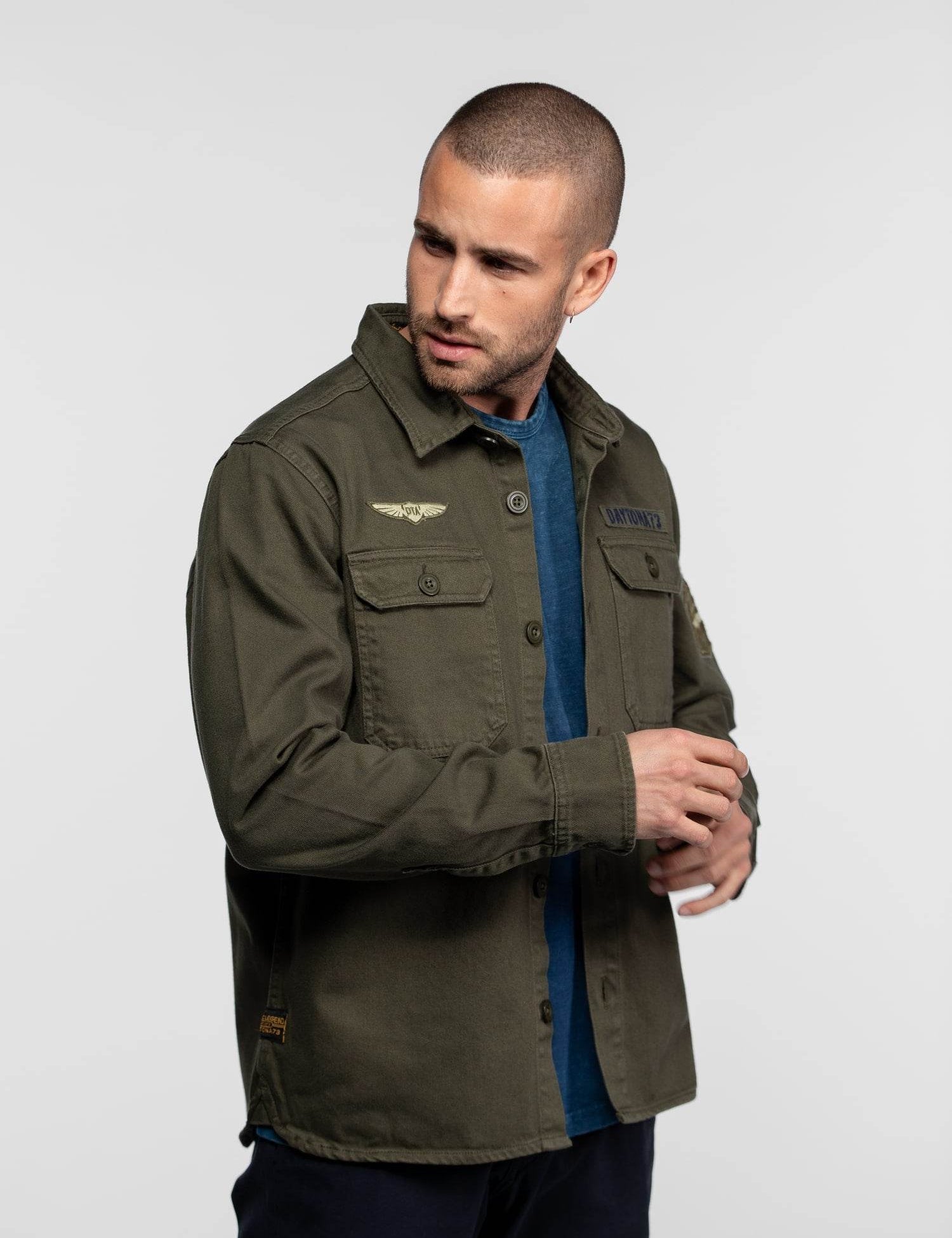 Man in green military-style jacket with patches, looking to the side, wearing a blue shirt underneath, against a plain background.