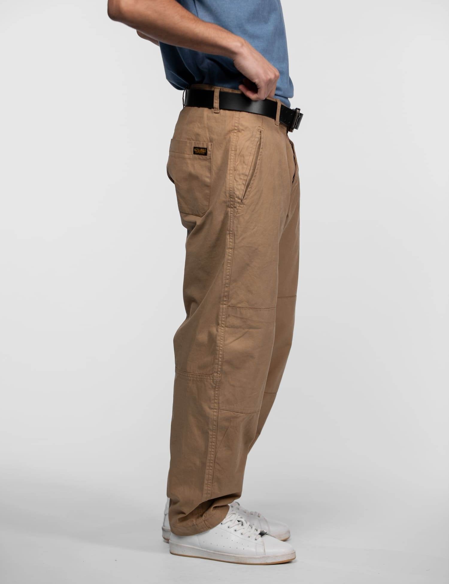 Man in blue shirt adjusts black belt on tan pants, wearing white sneakers, standing against a plain white background.