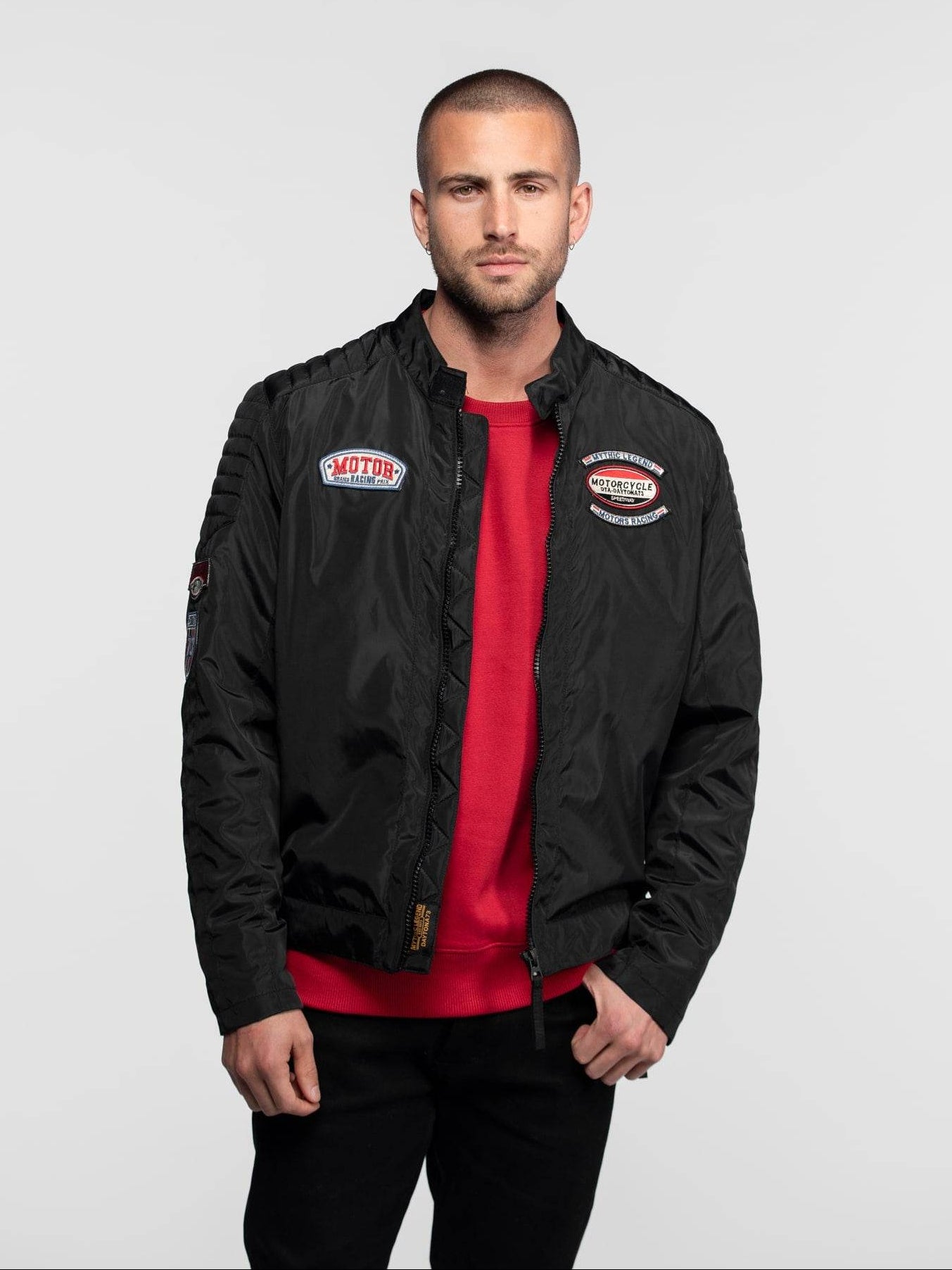 Homme portant un blouson noir avec écussons et pull rouge.