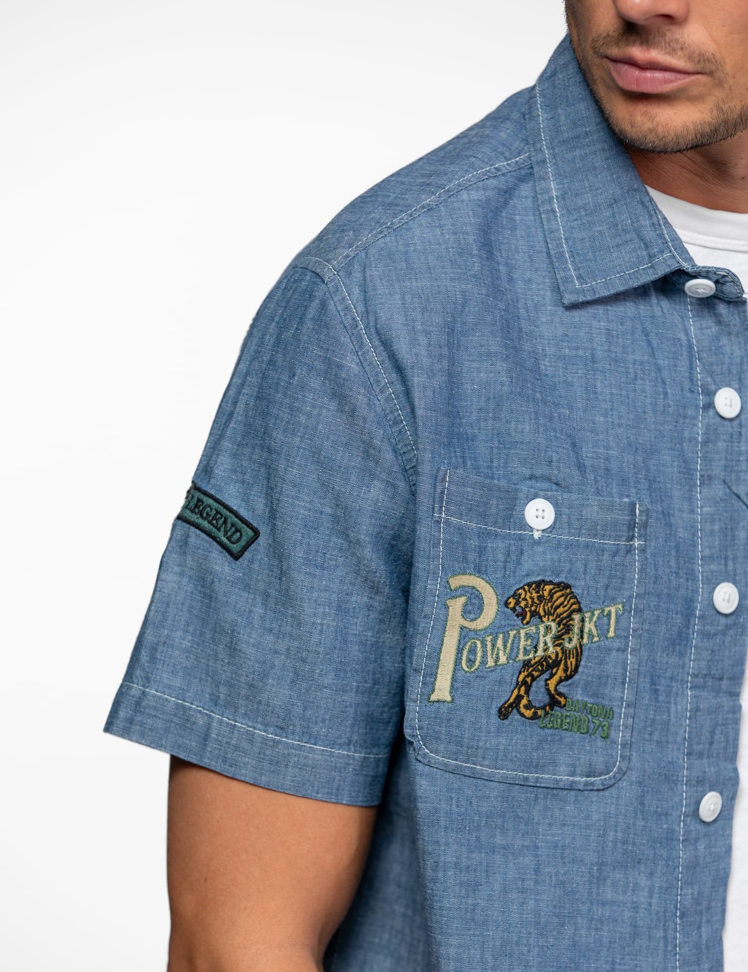 Man in a blue chambray shirt with "POWER JKT" and tiger embroidery on pocket, "LEGEND" patch on sleeve, white buttons.