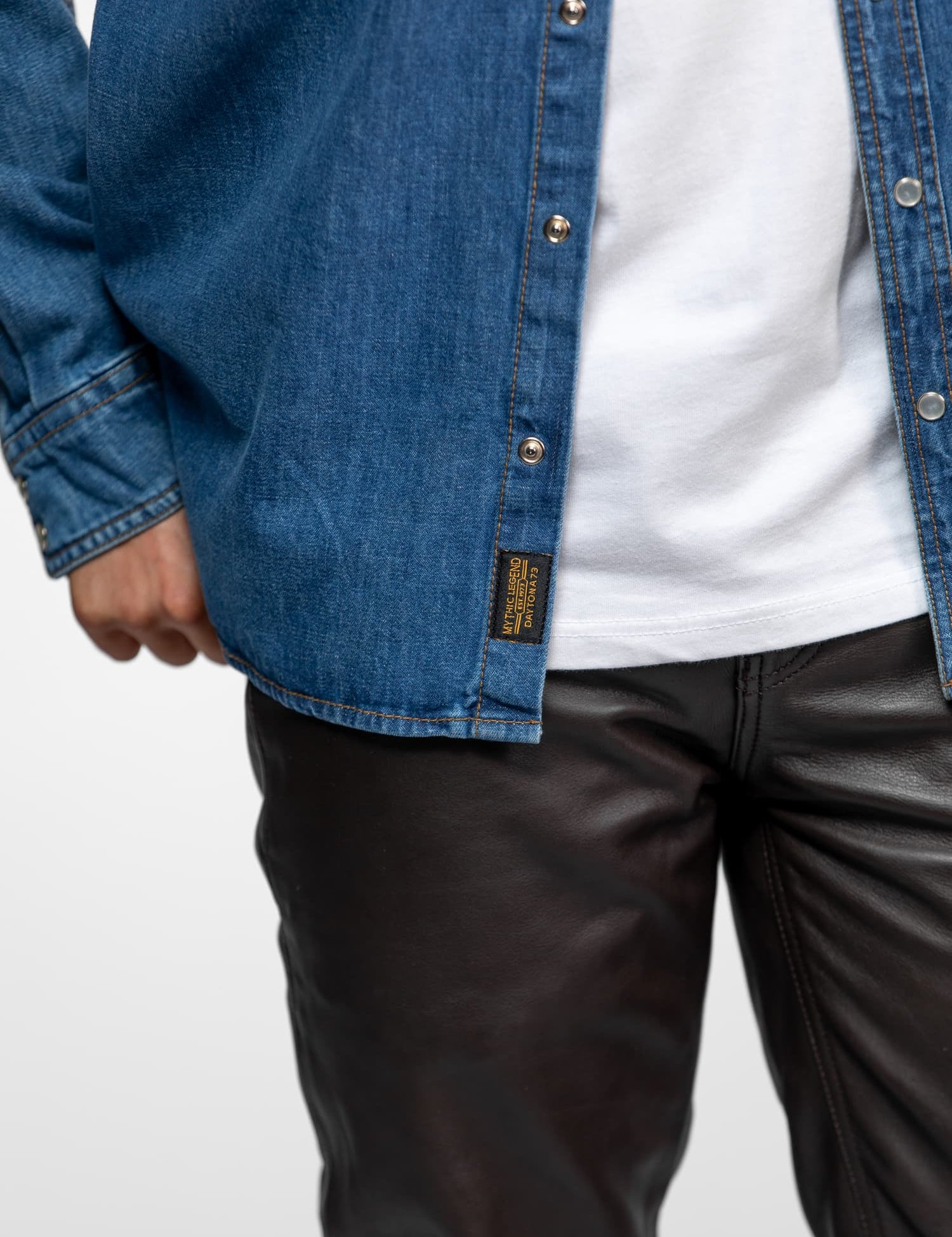 Person wearing a blue denim shirt with snap buttons over a white tee, paired with black leather pants, hand resting by side.