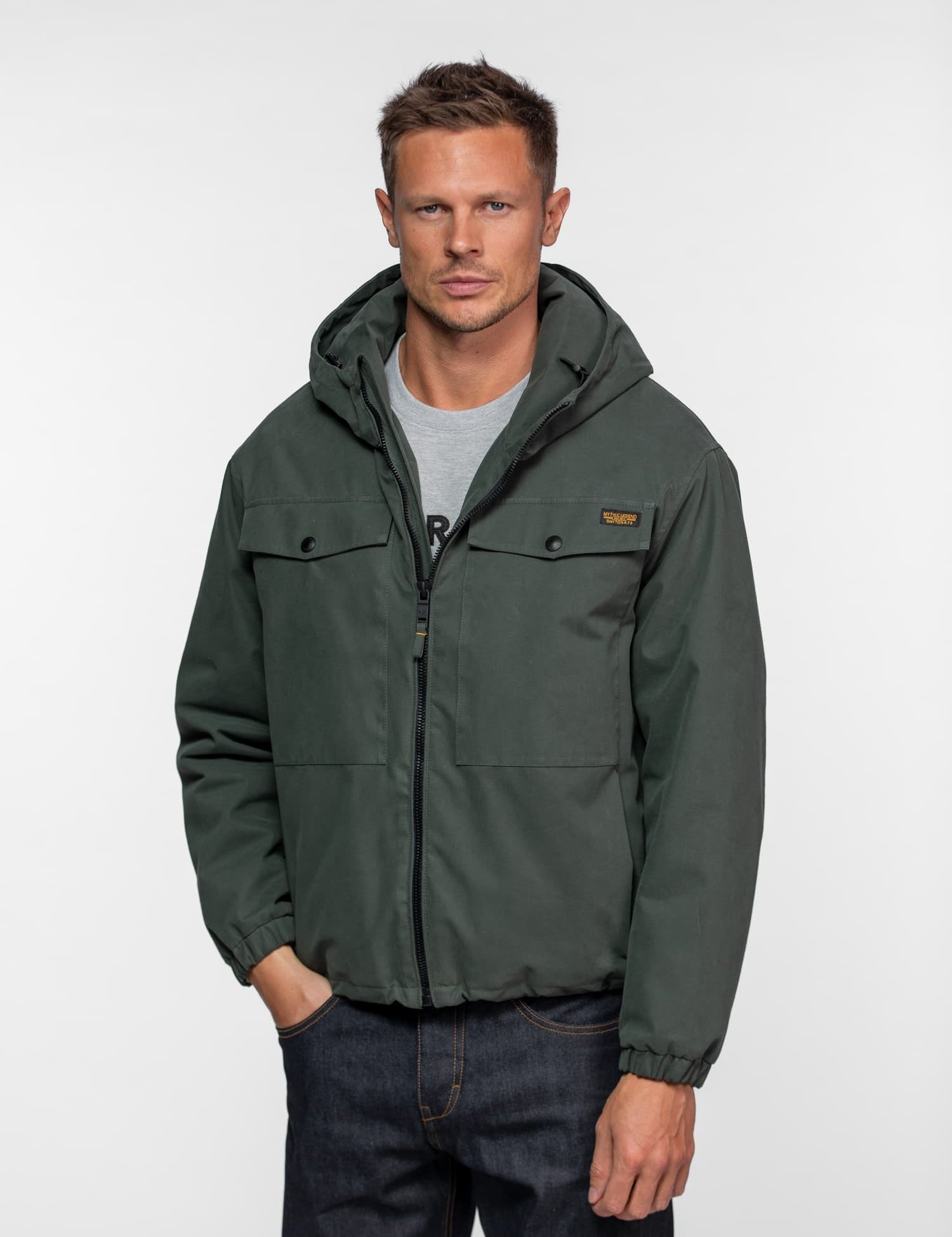 Man in a green hooded jacket and jeans stands against a plain background, hands in pockets, looking directly at the camera.