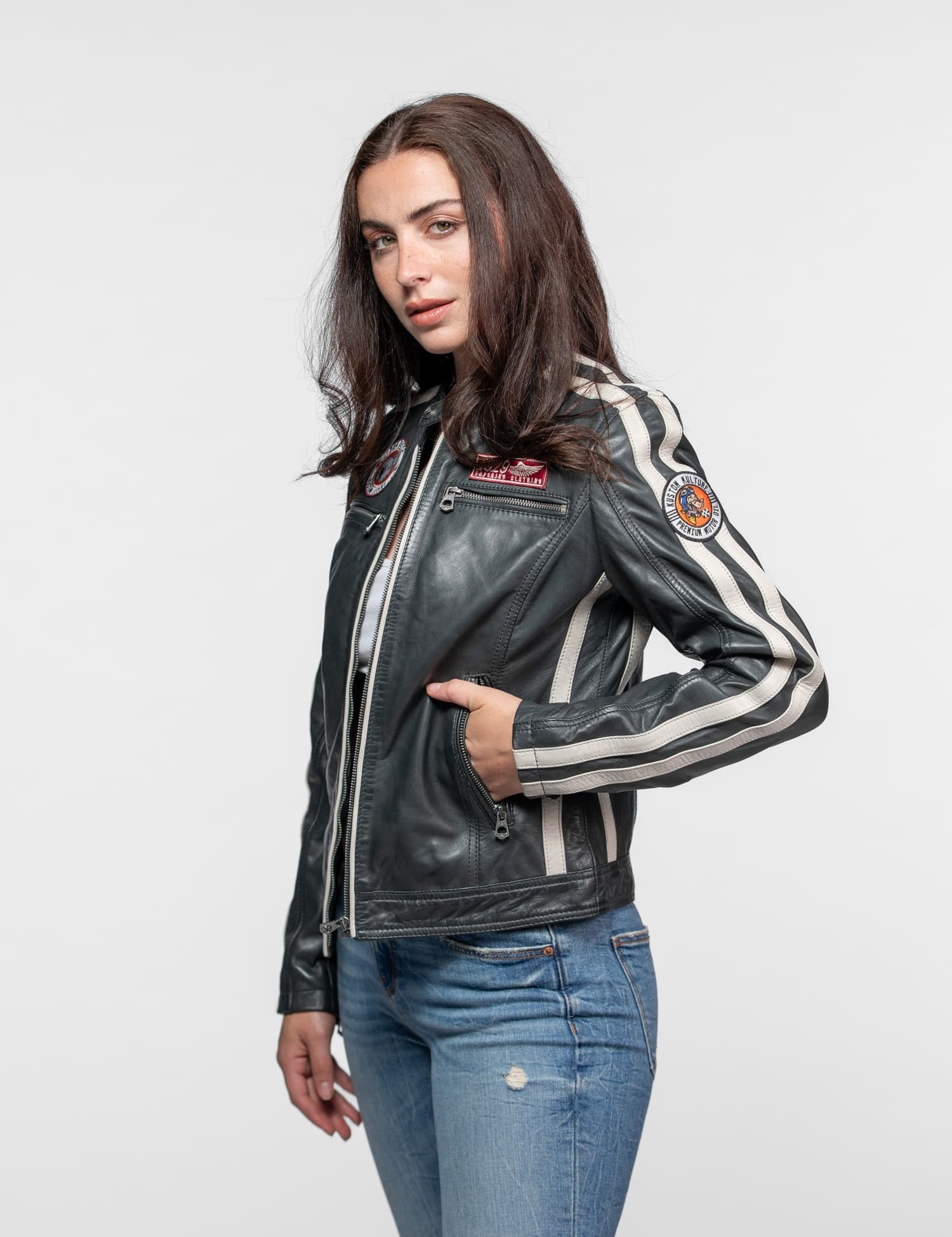 Woman in a black leather jacket with stripes and patches, hands in pockets, wearing blue jeans, standing against a plain background.