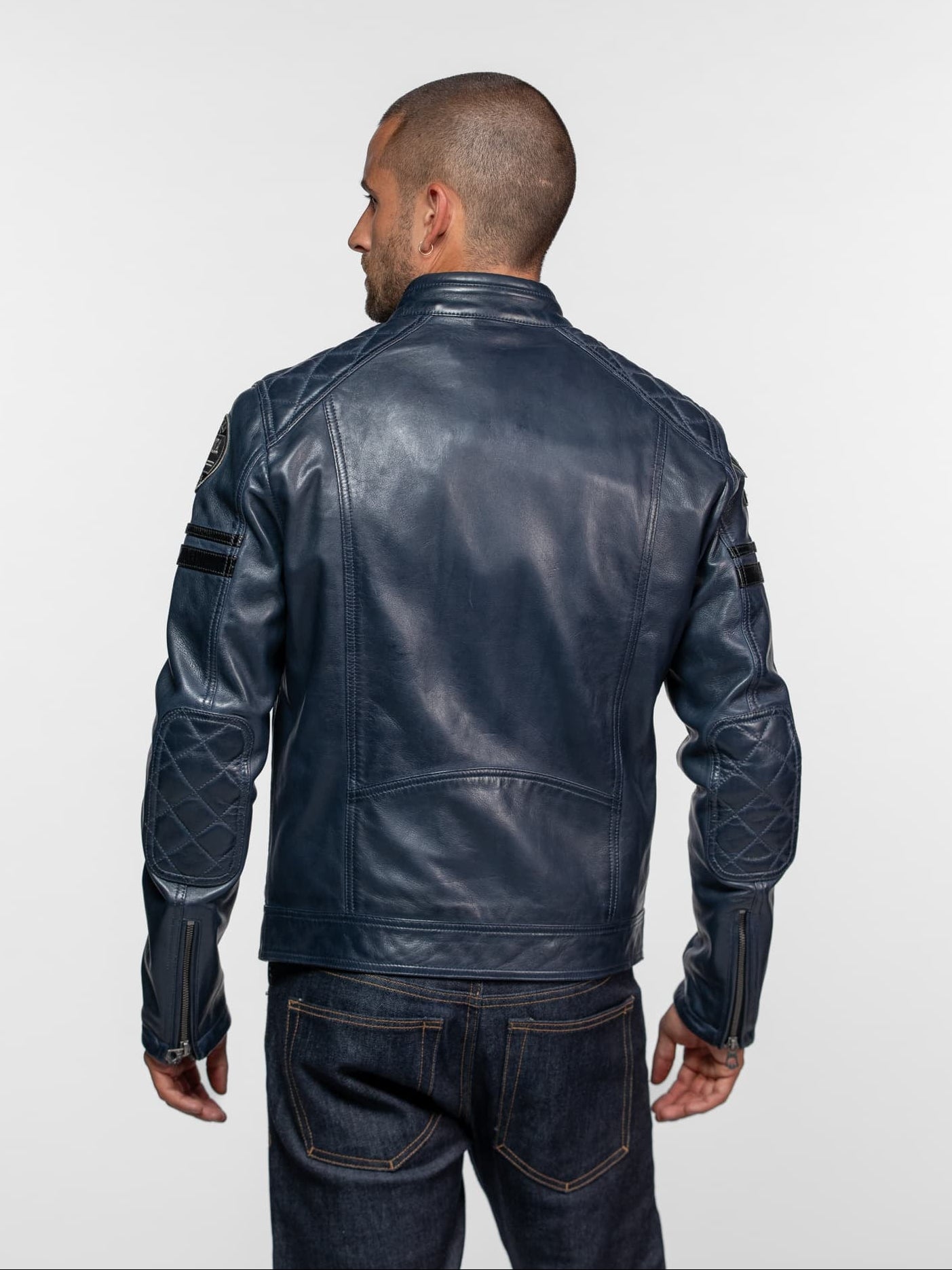 Man in a blue leather jacket with quilted elbows, viewed from behind, wearing dark jeans against a plain background.