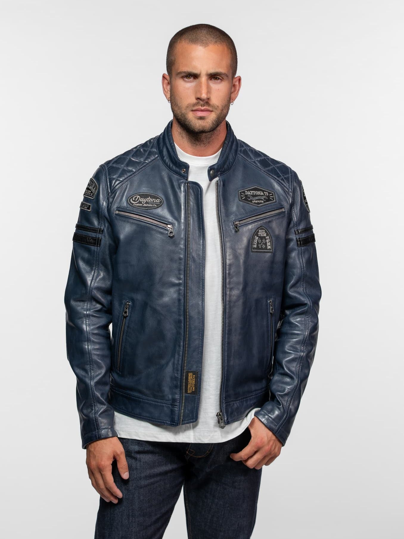 Man in a blue leather jacket with patches stands confidently against a plain background, wearing a white shirt and jeans.