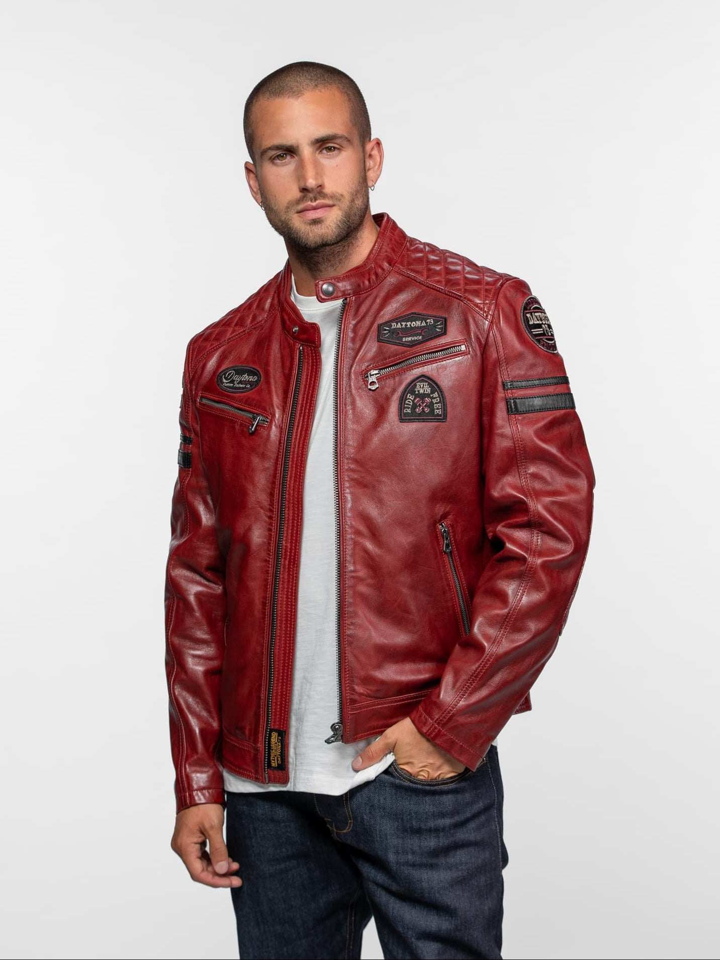 Man in a red leather jacket with patches, white shirt, and jeans, stands confidently against a plain background.