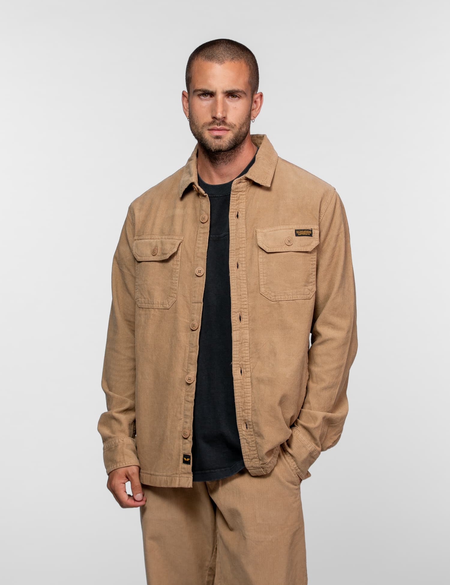Man in a beige corduroy jacket and pants, with a black shirt, stands against a plain background, hands in pockets, looking serious.