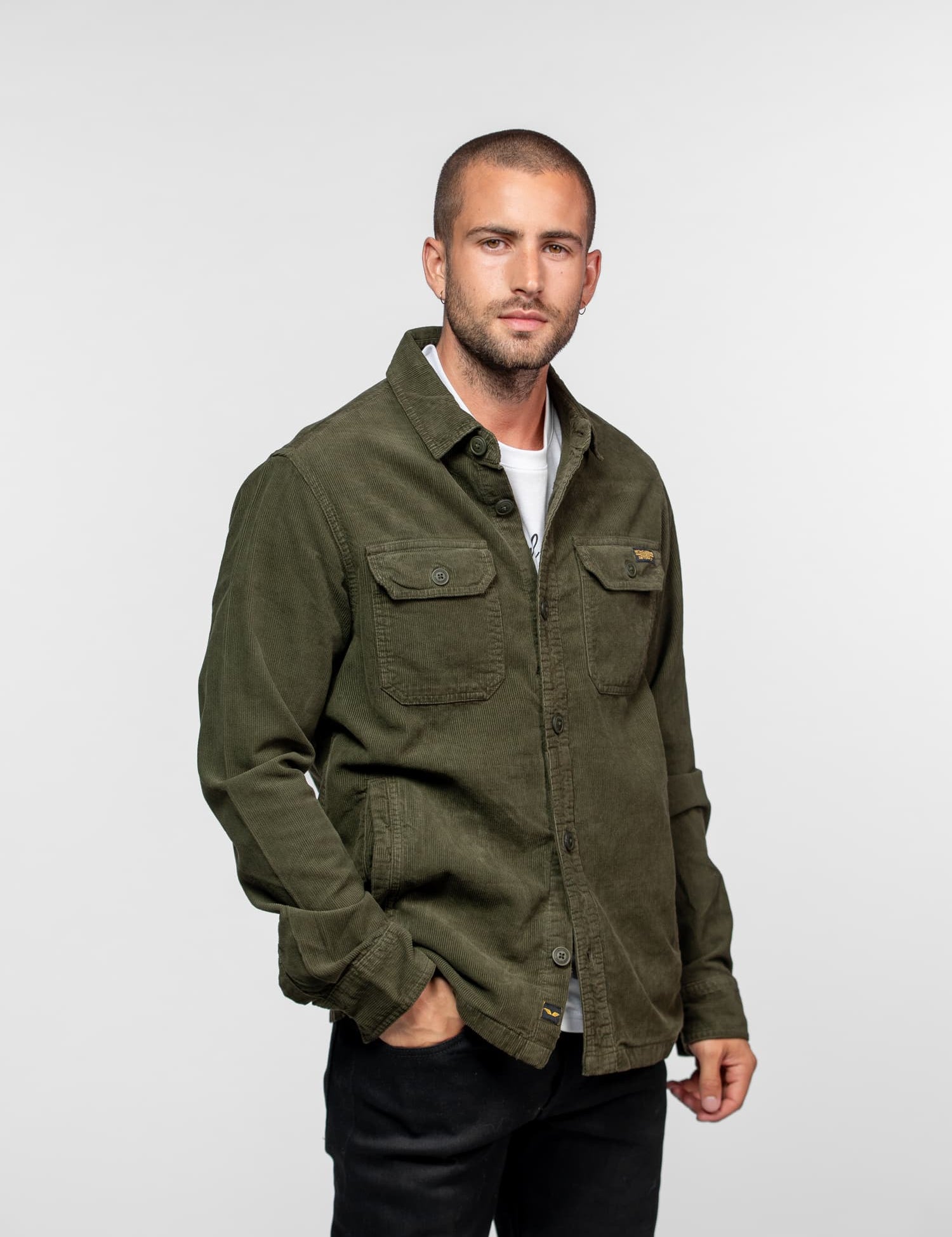 Man in a green jacket and black pants stands against a white background, looking confidently at the camera with hands in pockets.