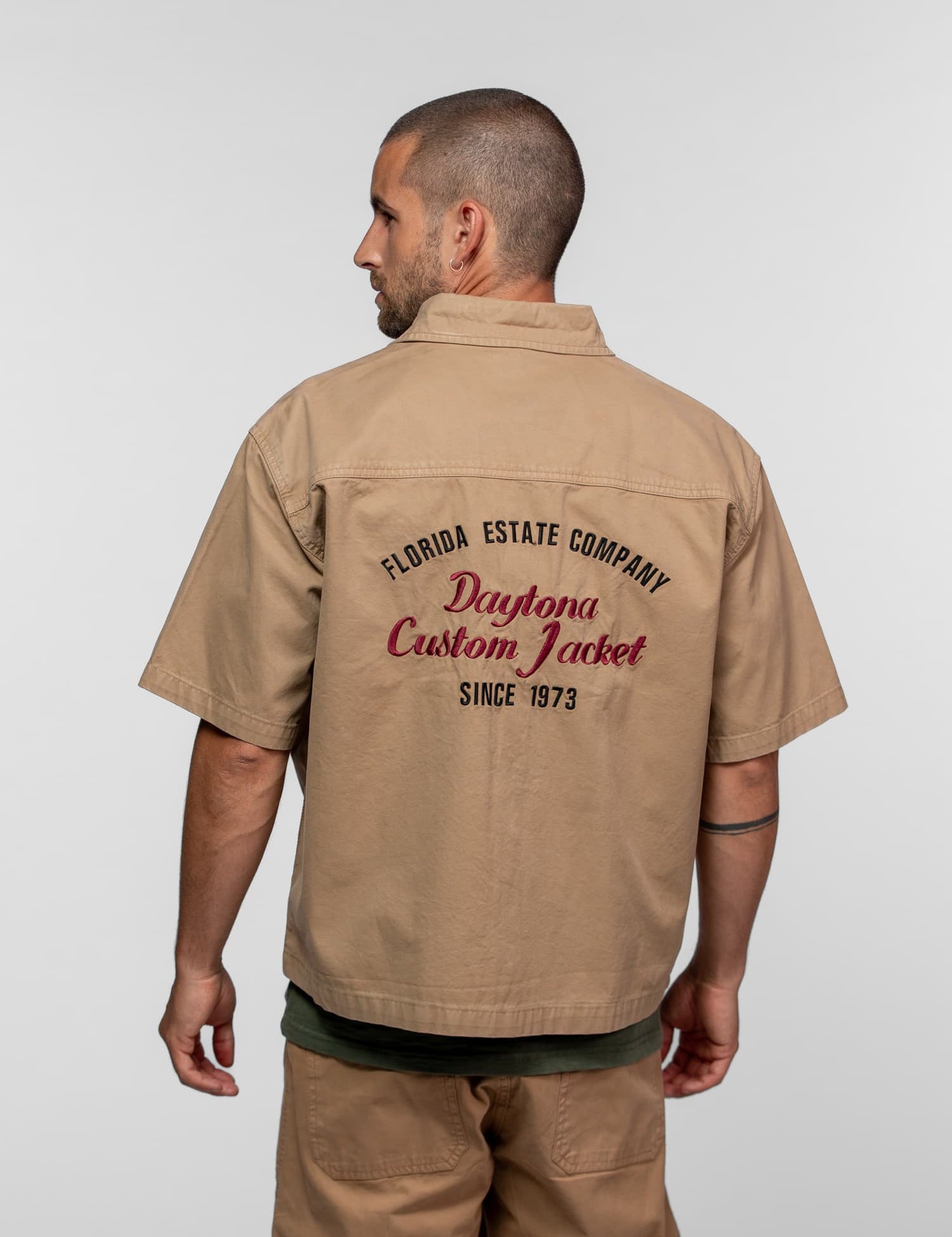 Man in beige shirt with "Florida Estate Company, Daytona Custom Jacket, Since 1973" text, facing left against a gray background.