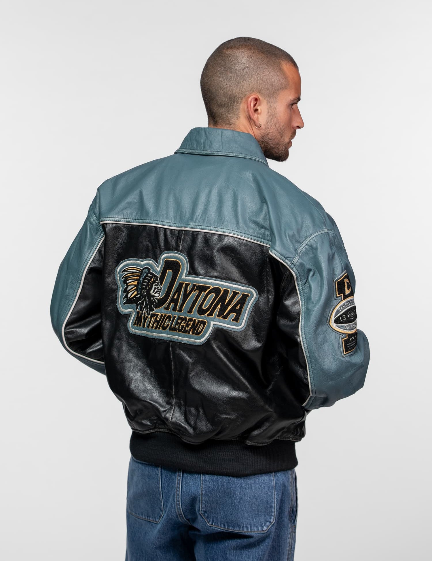 Man in blue jeans and leather jacket with "Daytona Mythic Legend" logo, facing sideways against a plain background.
