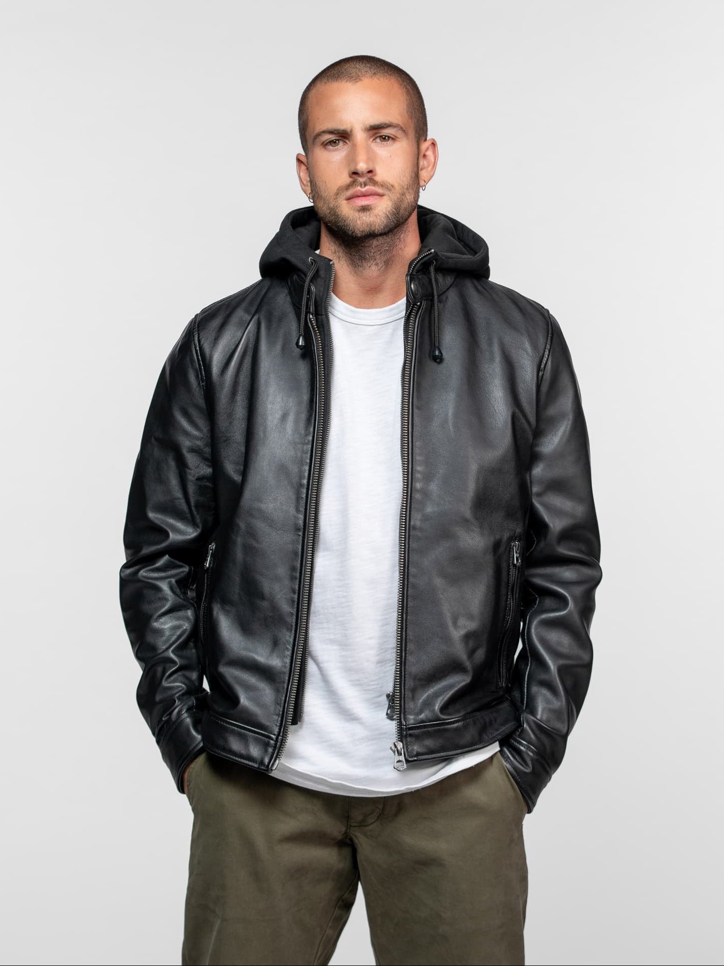 Man in a black leather jacket with a hood, white shirt, and khaki pants stands against a plain background, hands in pockets.