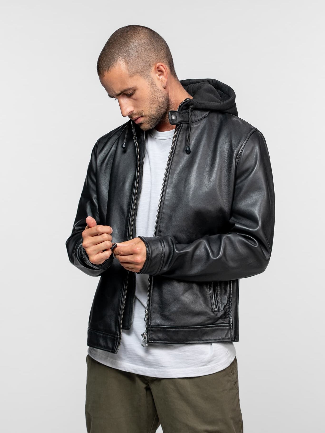 Man in a black leather jacket with a hood, white shirt, and green pants, looking down while adjusting his sleeve against a gray background.