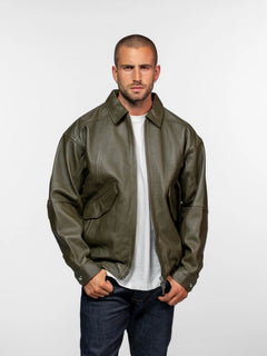 Man in olive leather jacket and jeans stands confidently against a plain white background, hands in pockets, looking forward.