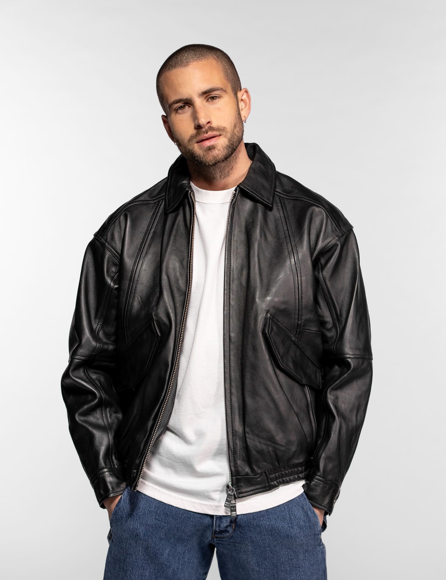 Un homme en blouson de cuir noir et jean se tient avec assurance sur un fond uni, les mains dans les poches, vêtu d'une chemise blanche.