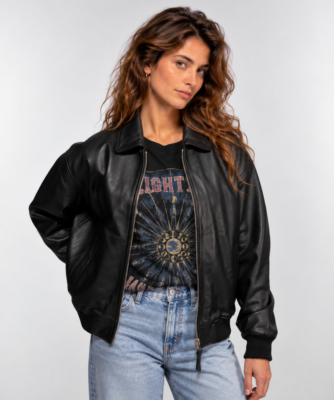 Femme en veste de cuir noire et t-shirt à motif, les mains dans les poches, pose avec assurance sur fond gris.