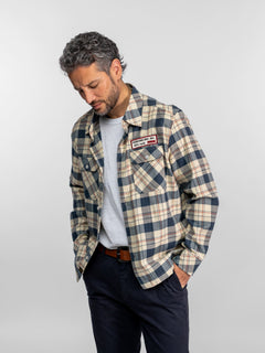 Homme en chemise à carreaux avec un écusson « Genuine Ind. Inc. Est. 1973 », regardant vers le bas, les mains dans les poches, sur fond uni.