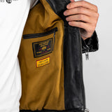 Une personne ouvre une veste en cuir noir révélant une doublure moutarde ornée d'un écusson Daytona de 1973 et d'une citation inspirante.