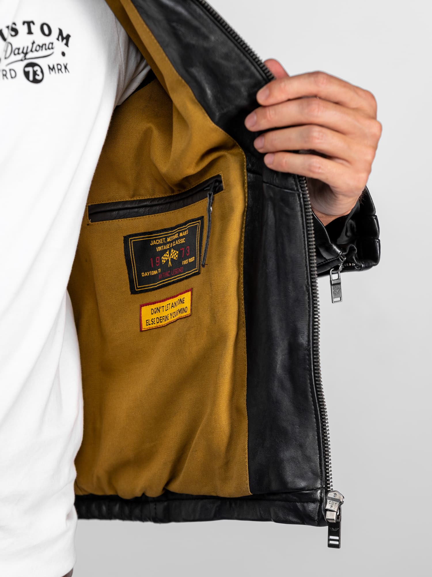 Une personne ouvre une veste en cuir noir révélant une doublure moutarde ornée d'un écusson Daytona de 1973 et d'une citation inspirante.