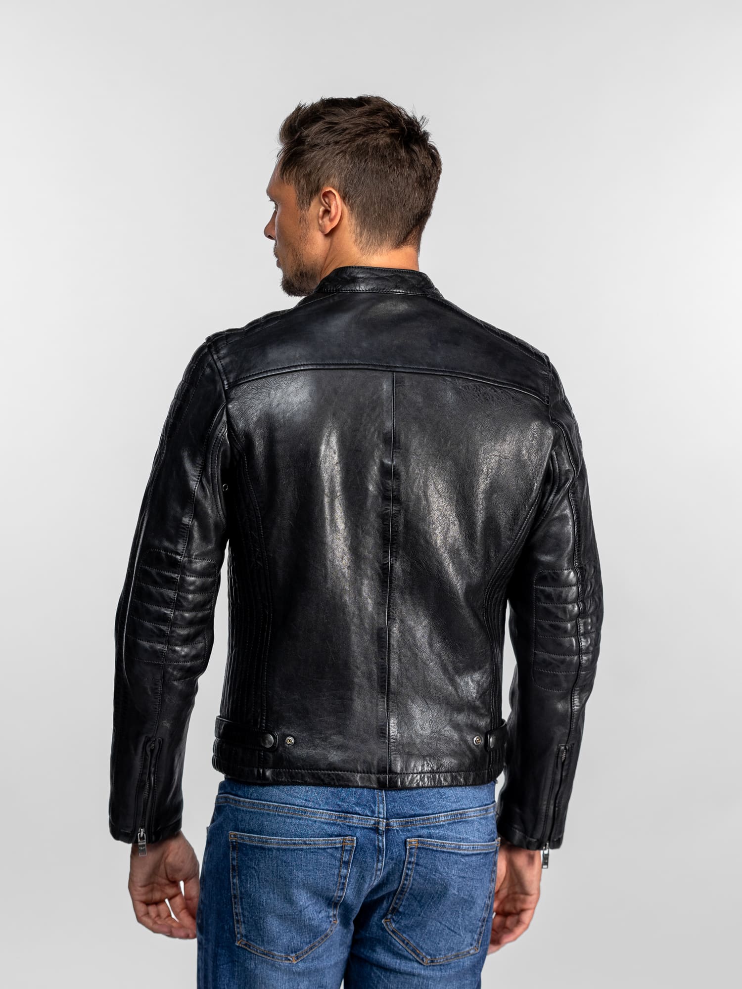 Un homme en veste de cuir noire et jean bleu, de dos, sur fond uni, mettant en valeur les détails du dos de sa veste.