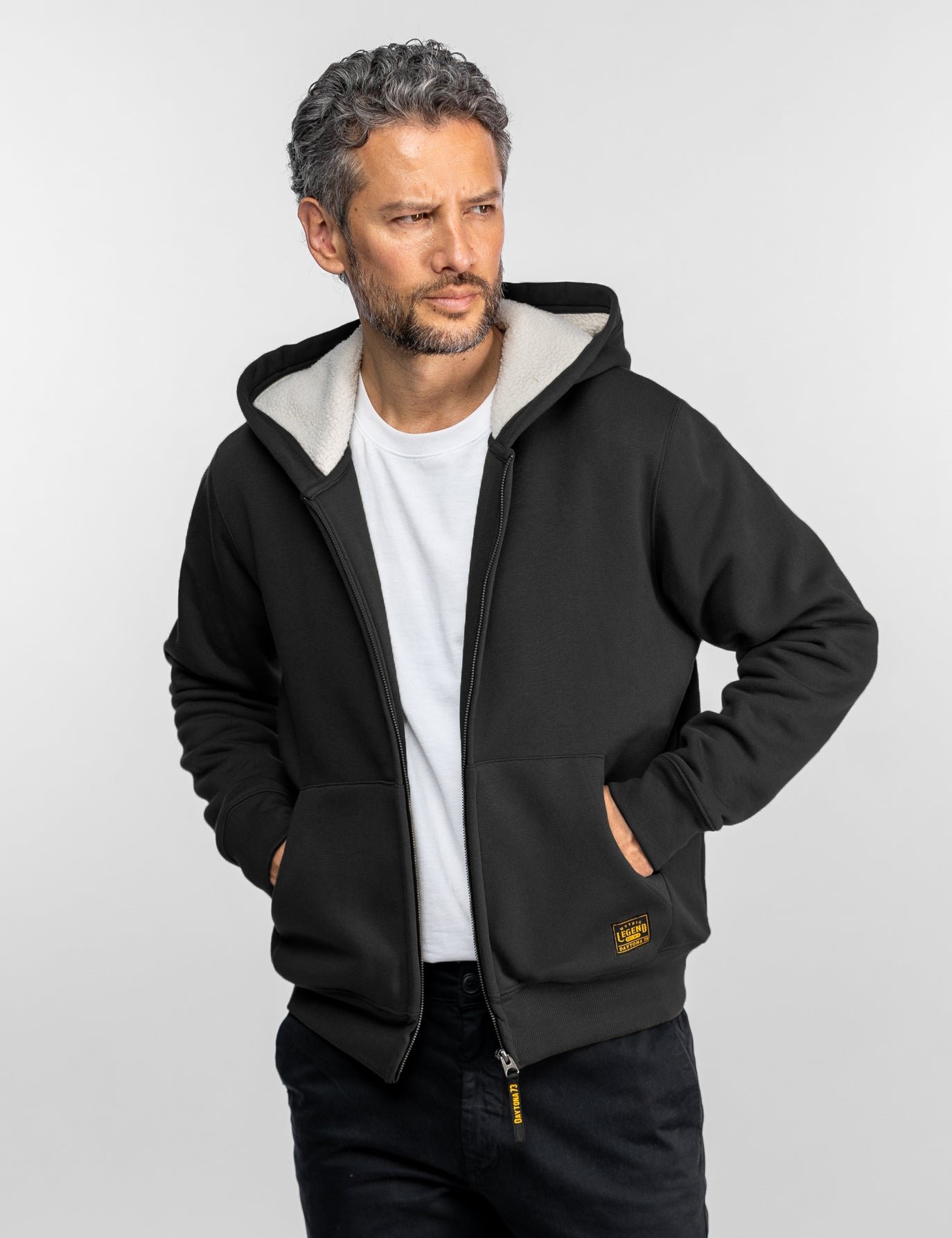 Un homme portant un sweat à capuche noir et une chemise blanche en dessous, regardant sur le côté, les mains dans les poches, sur un fond uni.