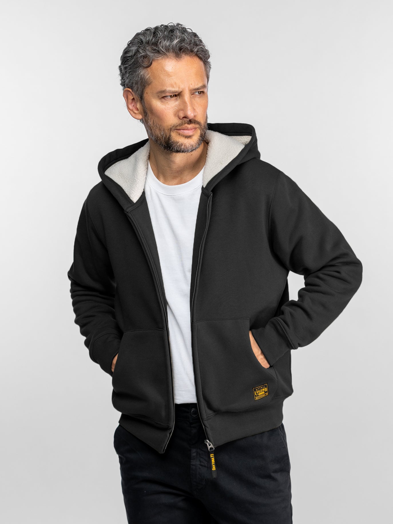 Un homme portant un sweat à capuche noir et une chemise blanche en dessous, regardant sur le côté, les mains dans les poches, sur un fond uni.