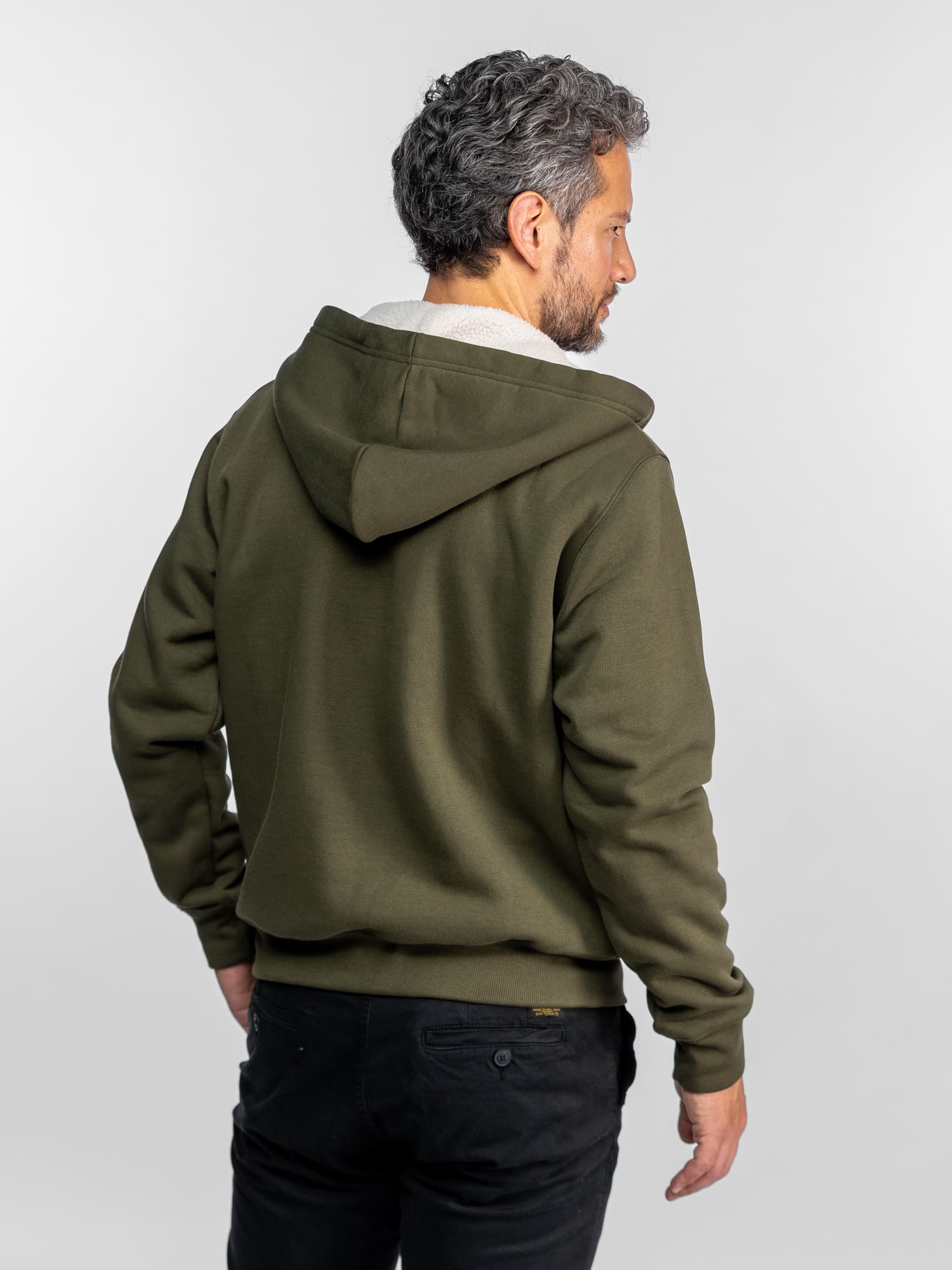 Homme aux cheveux gris, vêtu d'un sweat à capuche vert et d'un pantalon noir, vu de dos, sur fond blanc uni.