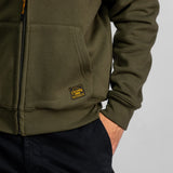 Personne portant un sweat à capuche olive avec un écusson « Legend », les mains dans les poches d'un pantalon noir, style décontracté.