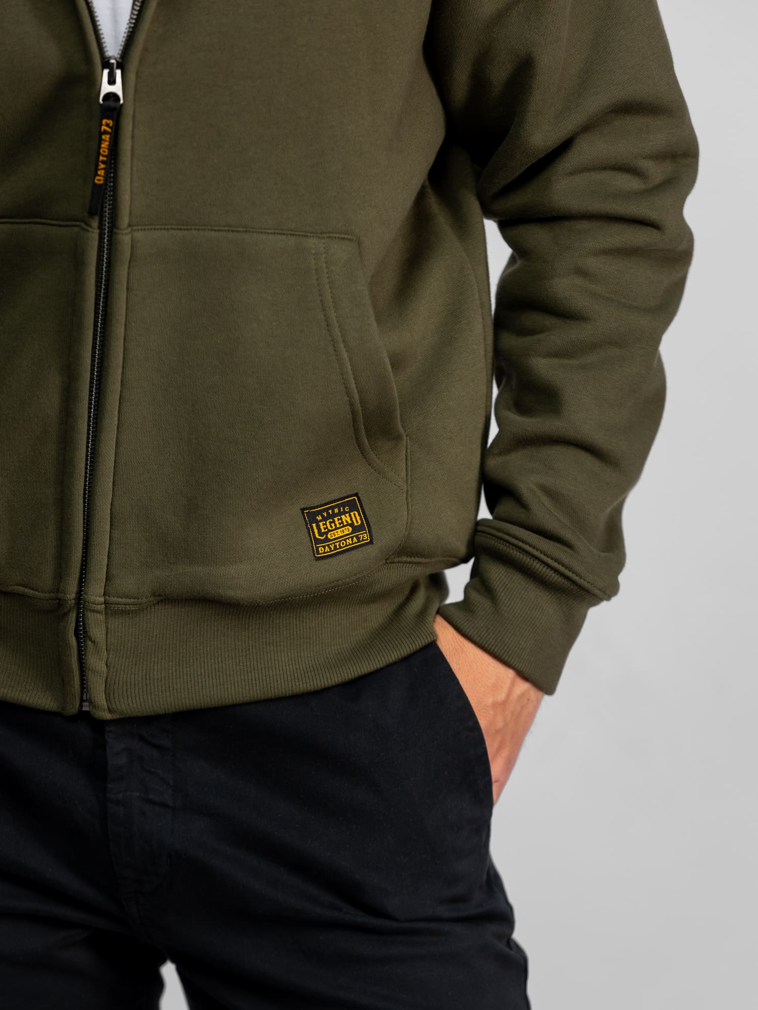 Personne portant un sweat à capuche olive avec un écusson « Legend », les mains dans les poches d'un pantalon noir, style décontracté.