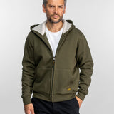 Un homme en sweat-shirt olive et chemise blanche se tient devant un fond uni, les mains dans les poches, regardant l'objectif avec assurance.