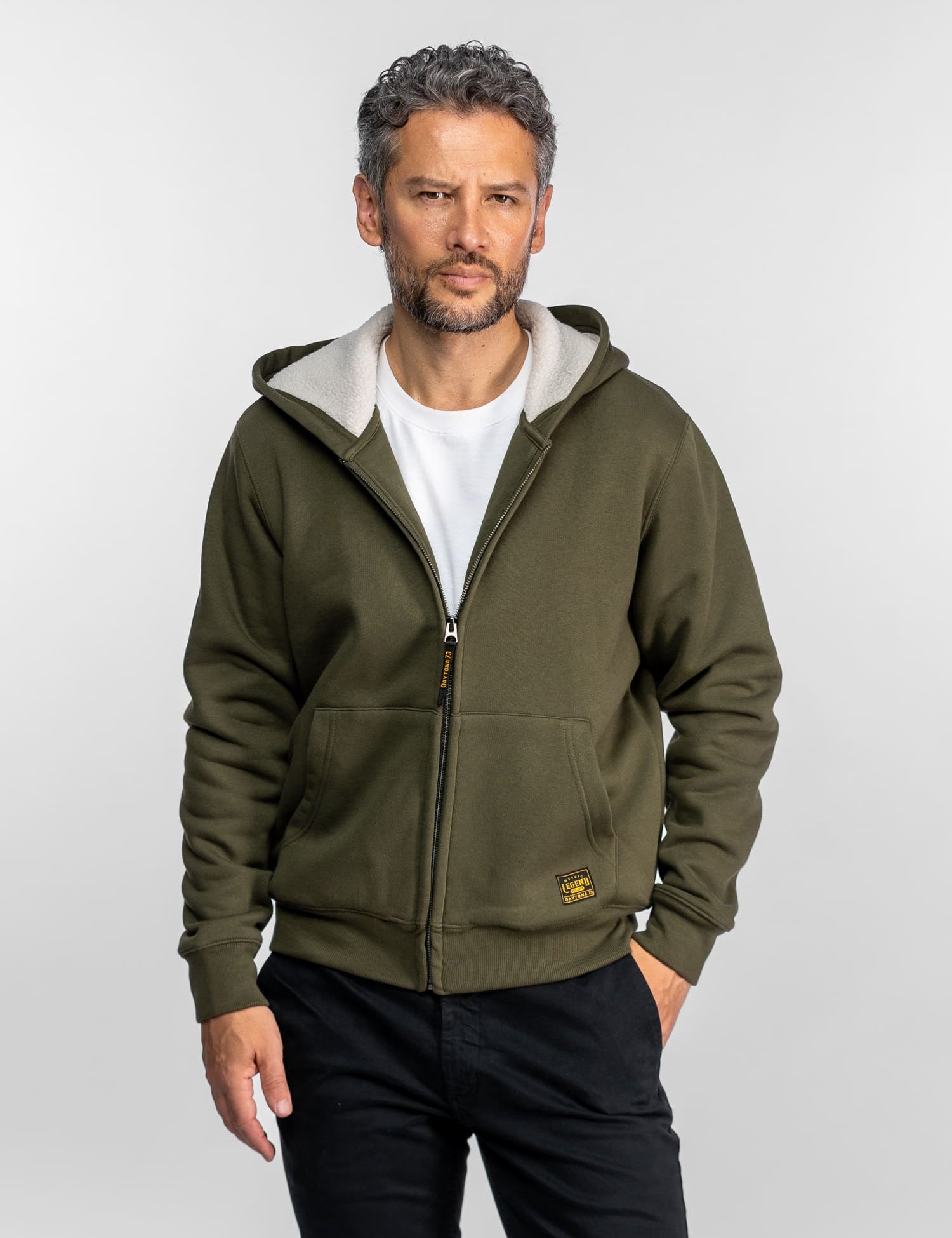Un homme en sweat-shirt olive et chemise blanche se tient devant un fond uni, les mains dans les poches, regardant l'objectif avec assurance.