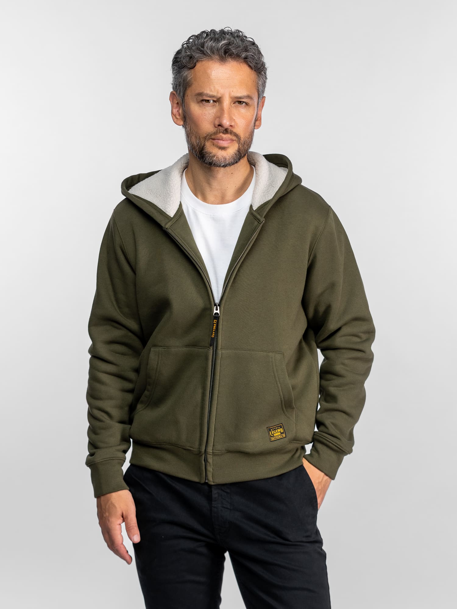 Un homme en sweat-shirt olive et chemise blanche se tient devant un fond uni, les mains dans les poches, regardant l'objectif avec assurance.