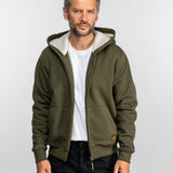 Un homme vêtu d'un sweat à capuche vert olive et d'une chemise blanche se tient avec assurance sur un fond uni, les mains dans les poches, regardant l'objectif.